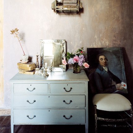 chest of drawers with flowers and a mirror, with a painting on a chair
